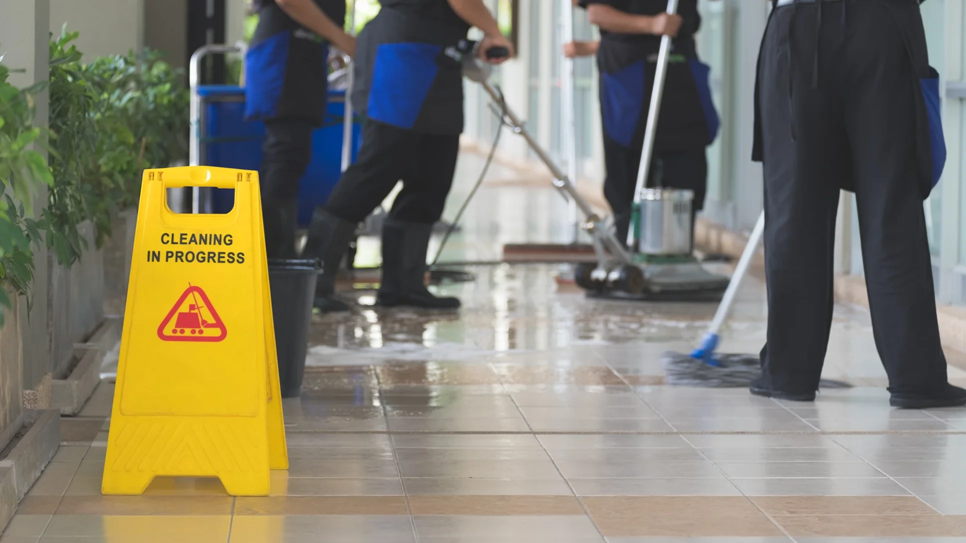 A group of people cleaning a floor, with a yellow warning sign indicating wet surfaces nearby.