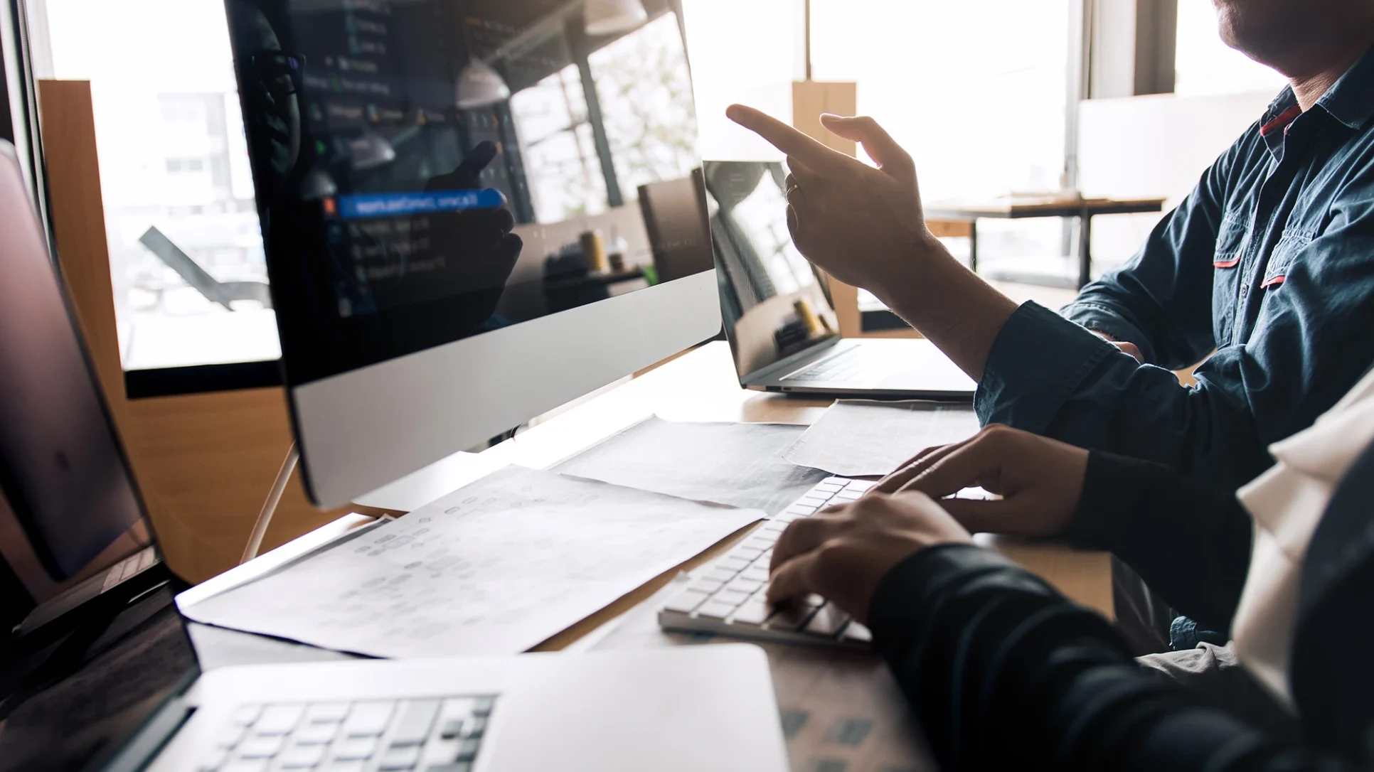 Two people collaborating on a project while working on a computer in front of a large monitor.