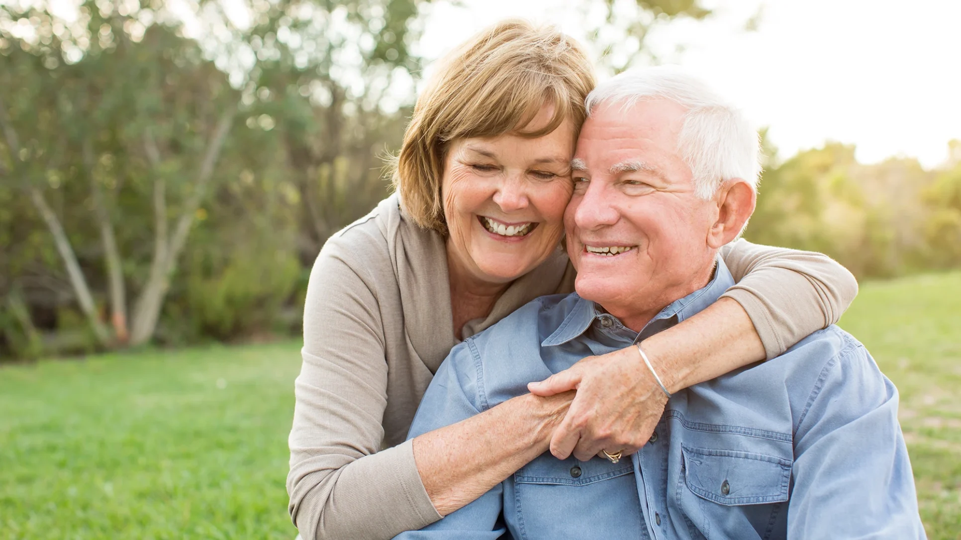 An older couple sits together on the grass in a park, enjoying a sunny day and each other's company.